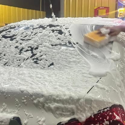 มือถือฟองน้ำเช็ดโฟมล้างรถบนรถสีขาวที่ P Selfwash (Hand applying a sponge to the foam-covered hood of a white car at P Selfwash)