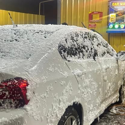 รถสีขาวเคลือบโฟมเต็มคันที่ช่อง P Selfwash พร้อมตู้หยอดเหรียญ CAR WASH ในพื้นหลัง (White car fully covered in cleaning foam at P Selfwash bay with the CAR WASH coin terminal visible behind)